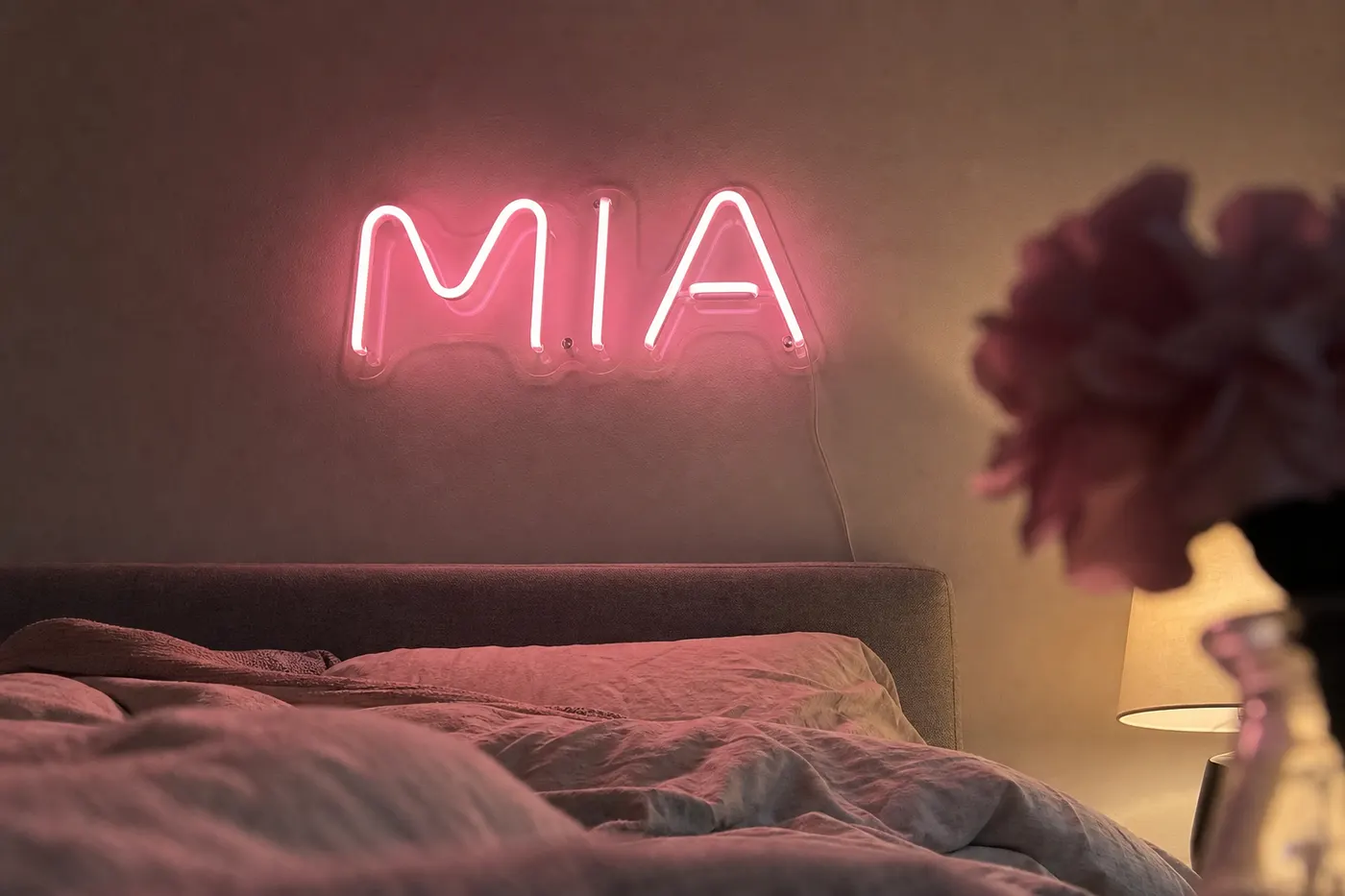 Pink MIA LED neon name sign above a bed with blurred bedding and flower